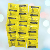 A cluster of bright yellow b Condoms Snugger Fit (49mm) packaging, each labeled for quality assurance and featuring the bCondoms logo and branding. The ultra-smooth natural rubber latex packaging is arranged in a slightly overlapping pattern on a light background with bokeh light effects.